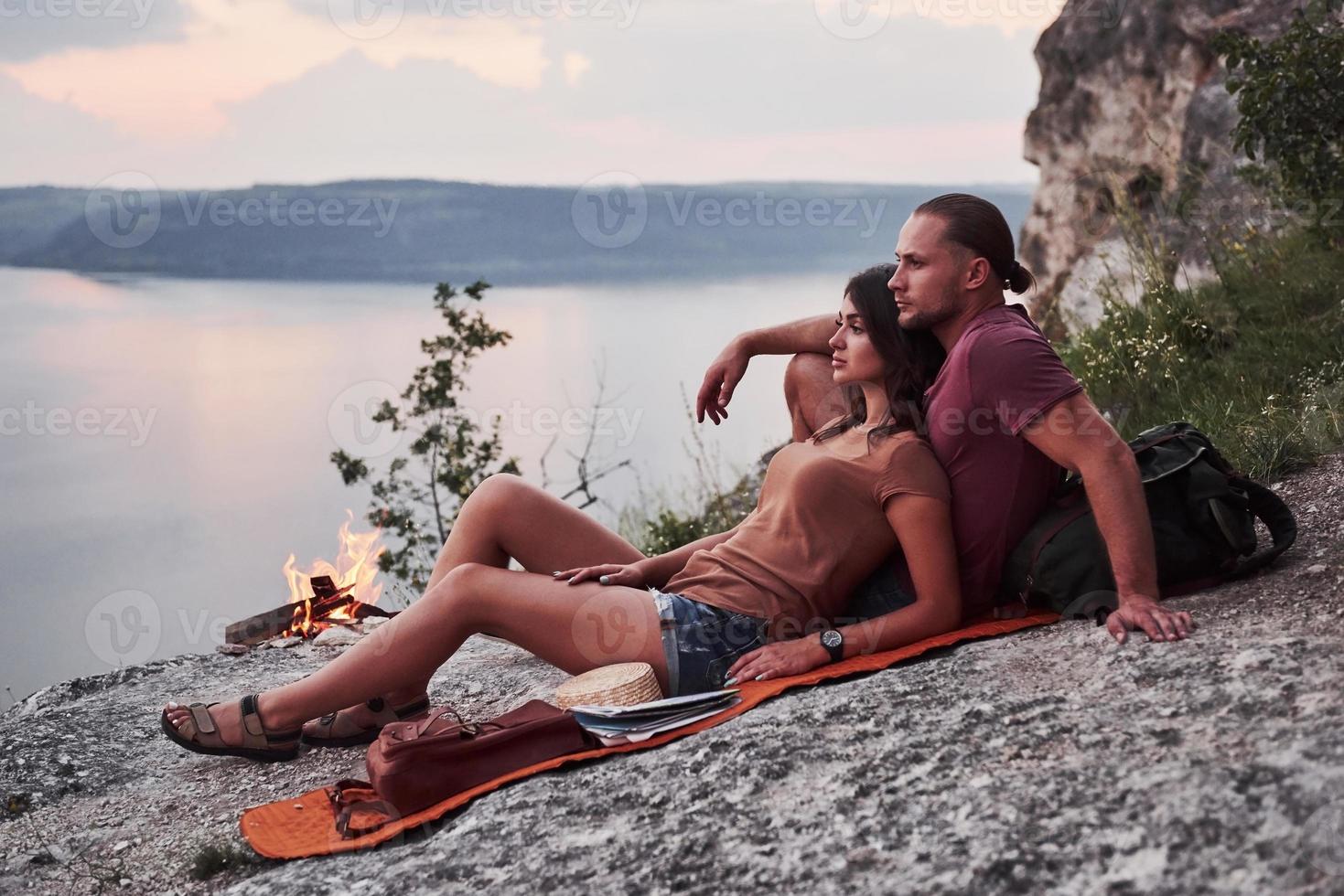 pareja abrazándose con mochila sentado cerca del fuego en la cima de la montaña disfrutando de la vista de la costa, un río o un lago. viajando a lo largo de las montañas y la costa, la libertad y el concepto de estilo de vida activo foto