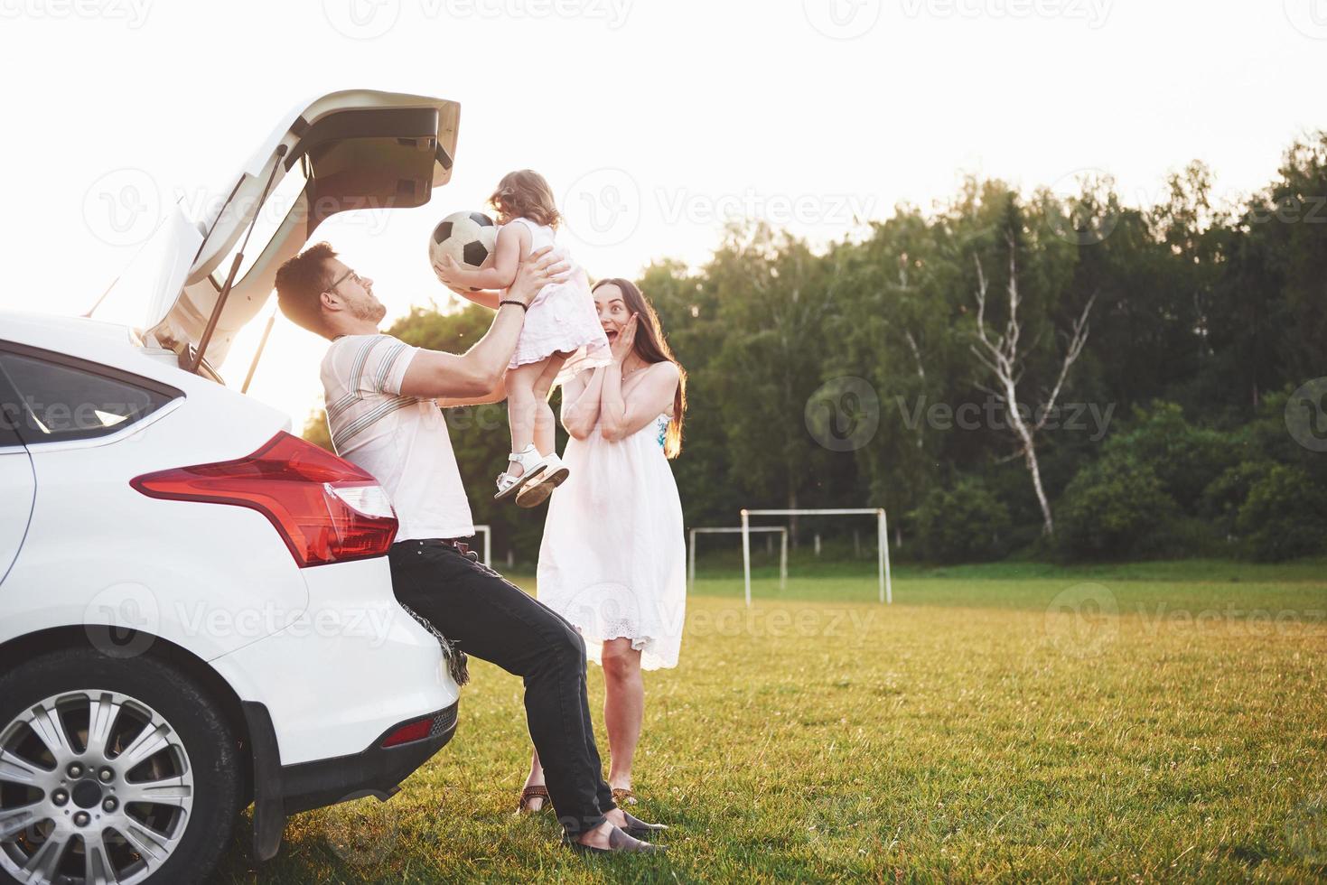 Pretty young married couple and their daughter are resting in the nature. The mother father and little girl are sitting on open car boot photo