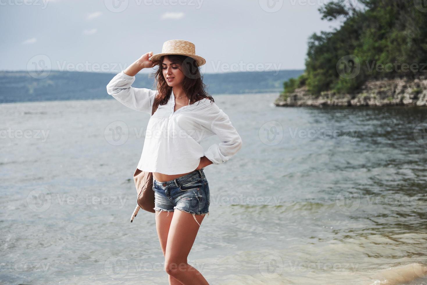 A beautiful young girl in a hat and with a backpack playfully walks by the water. A warm summer day is a great time for adventure and adventure in nature photo