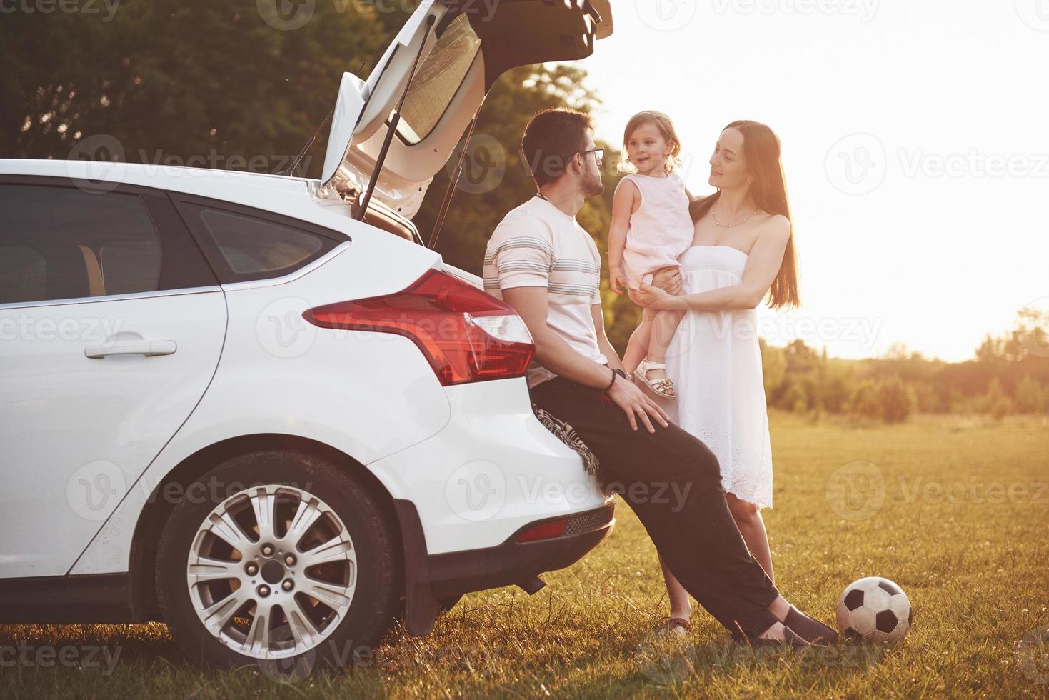 Pretty young married couple and their daughter are resting in the nature. The mother father and little girl are sitting on open car boot photo