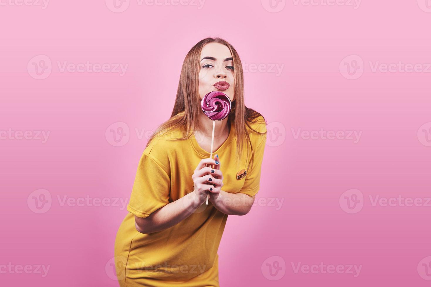 retrato divertido lindo de la muchacha que sostiene la piruleta que sonríe en fondo rosado. hermosa niña caucásica multicultural sonriendo feliz foto