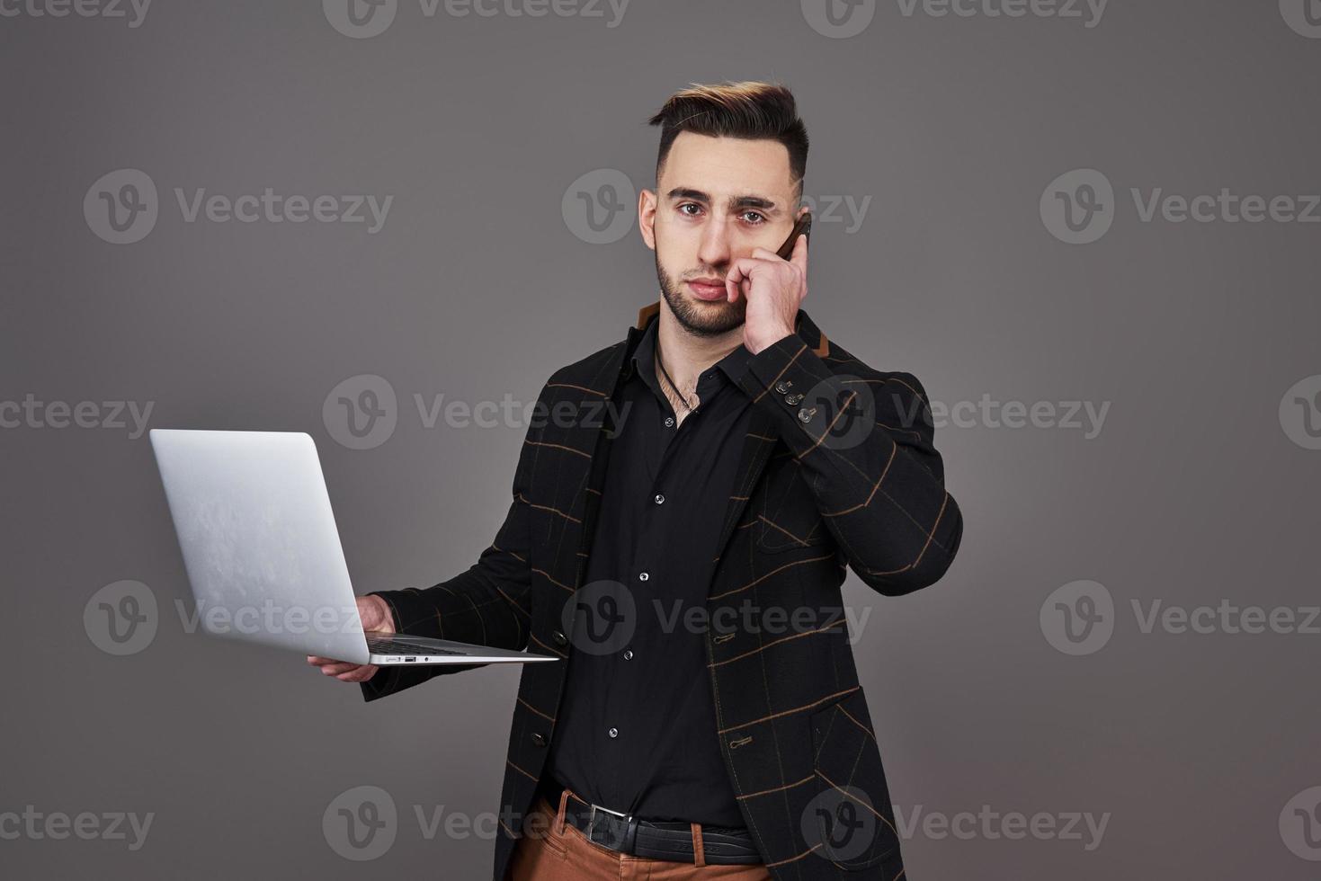 Busy bearded man in business clothes talking by smartphone and using laptop computer while holding cup of coffee in hand over gray background photo