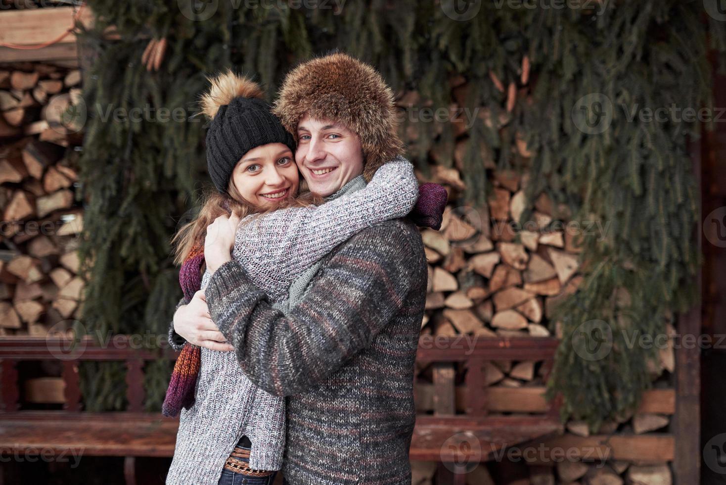 christmas and couple concept - smiling man and woman in hats and scarf hugging over wooden country house and snow background photo