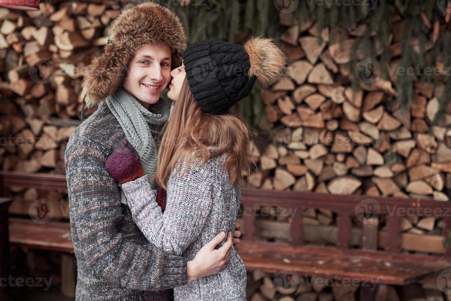 christmas and couple concept - smiling man and woman in hats and scarf hugging over wooden country house and snow background photo