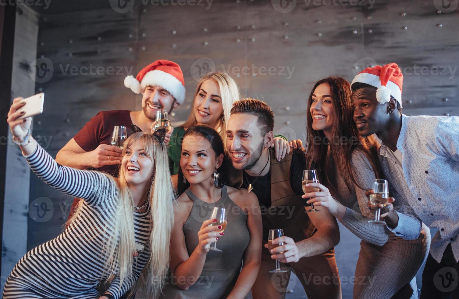 Agrupe a hermosos jóvenes haciendo selfie en la fiesta de año nuevo, mejores amigos niñas y niños divirtiéndose juntos, posando personas de estilo de vida emocional. sombreros de santa y copas de champán en sus manos foto