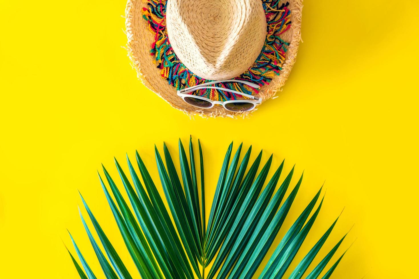 sombrero de paja y gafas de sol con hojas de palmera verde sobre un fondo amarillo brillante. concepto de verano, viajes a la playa. foto