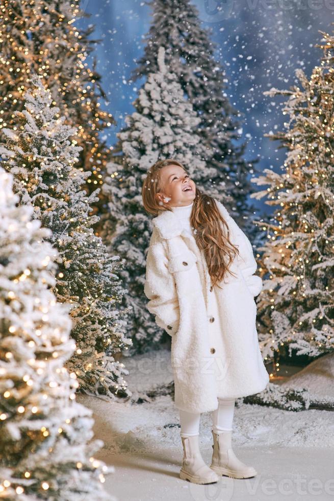 cute caucasian blonde girl in white eco fur coat walking in winter christmas forest with lights. New year, fairy tale concept photo
