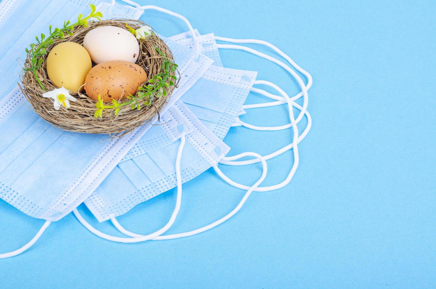 nido con huevos teñidos de colores brillantes con máscaras médicas sobre fondo azul, espacio para texto. vacaciones de pascua durante la pandemia de coronavirus foto