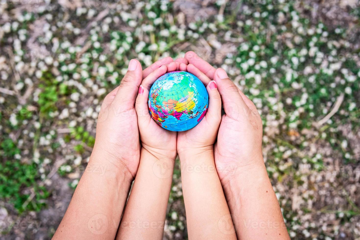 primer plano la mano de la madre abraza la mano de la hija abrazando nuestro globo en nuestras palmas sobre fondo de tierra con hierba verde y flores blancas. desenfoque de fondo. concepto para ayudar a cuidar nuestro mundo. foto