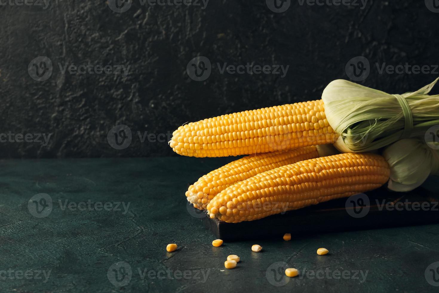 Wooden board with fresh corn cobs on dark background photo