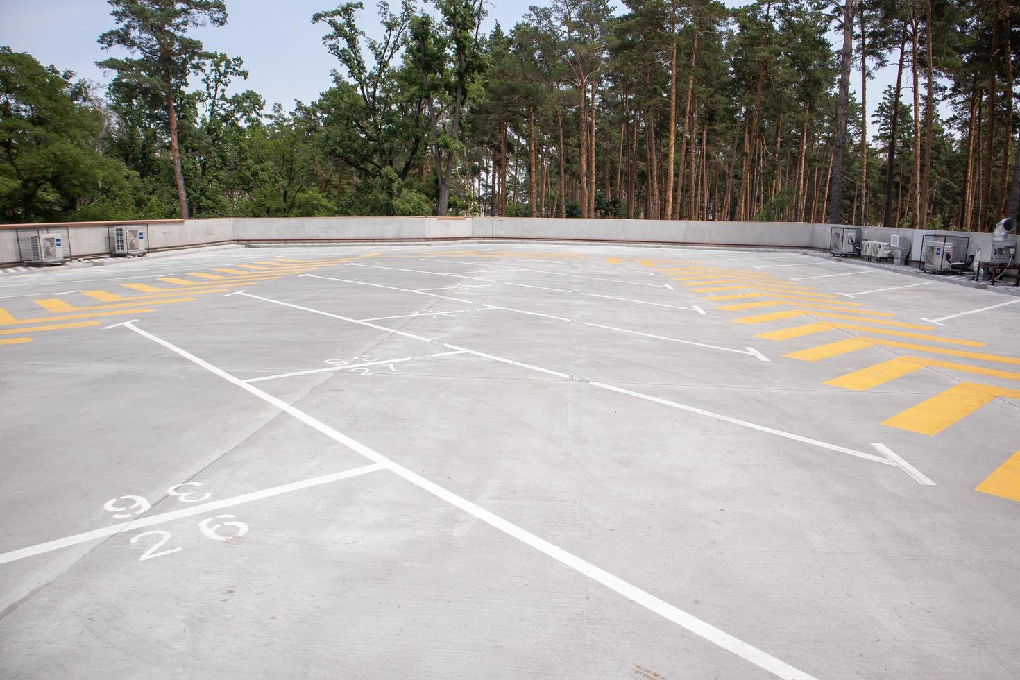 Empty rooftop parking. Directional traffic arrows and parking spots are marked with white and yellow paint on the asphalt. Parking space without cars with city skyline and park with trees. photo