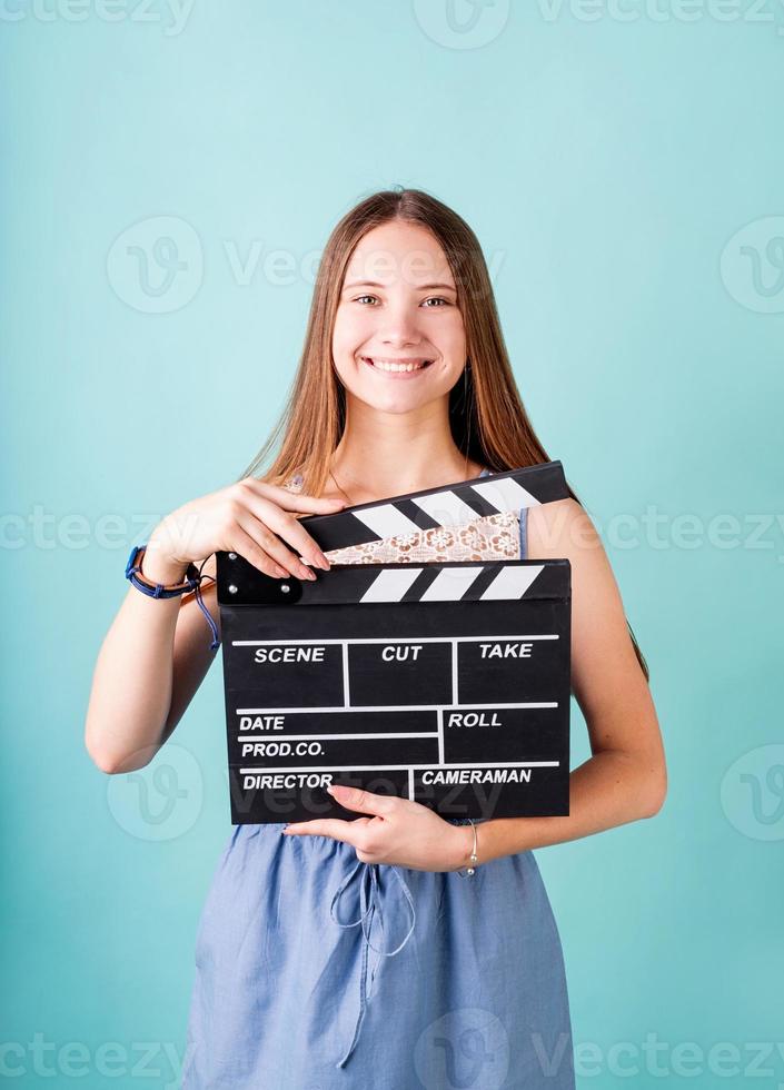 Happy smiling teenager girl in a blue dress holding a clapper board isolated on blue photo