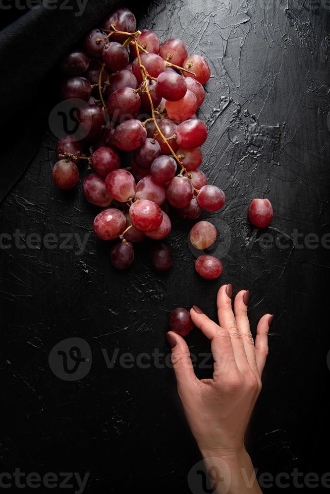 Woman hand holding a grape from a bunch of red grapes top view on dark background photo