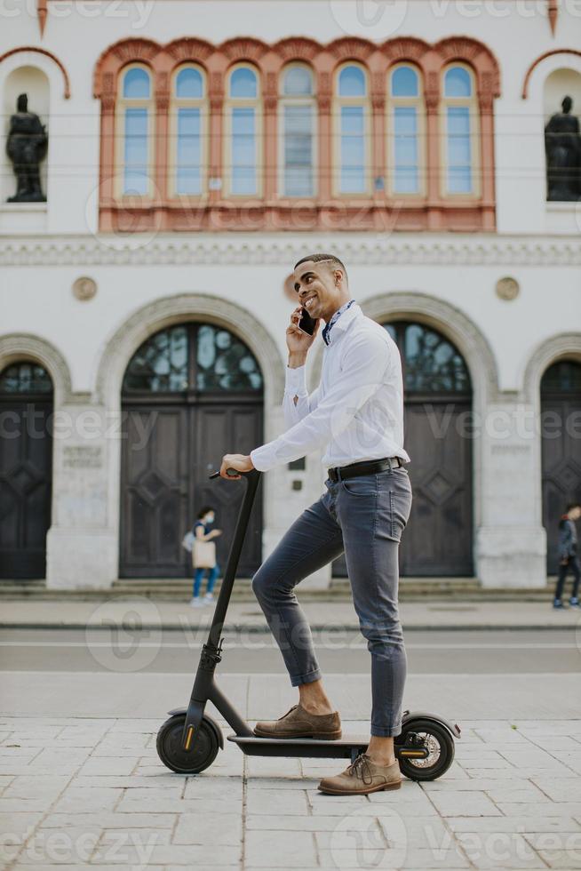 Joven afroamericano mediante teléfono móvil mientras está de pie con scooter eléctrico en una calle foto