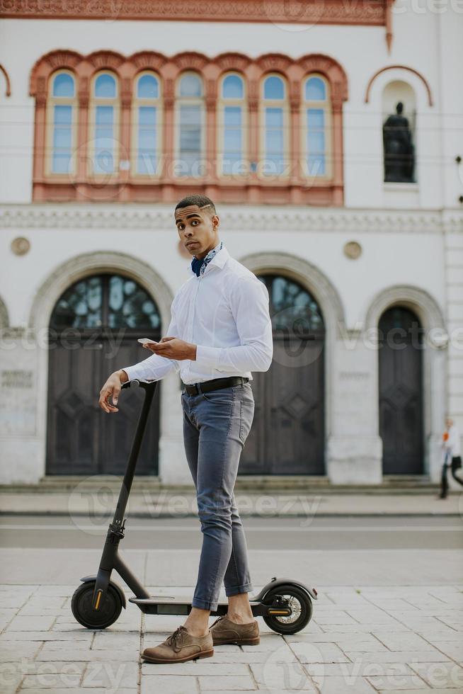 Joven afroamericano mediante teléfono móvil mientras está de pie con scooter eléctrico en una calle foto