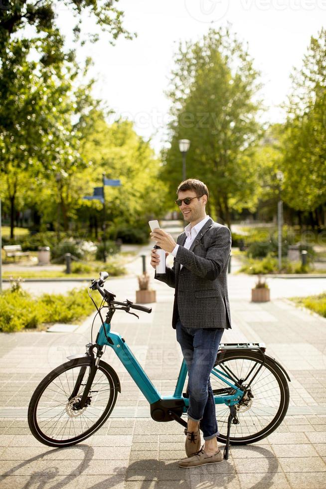 Young businessman using mobile phone by the ebike with takeaway coffee cup photo