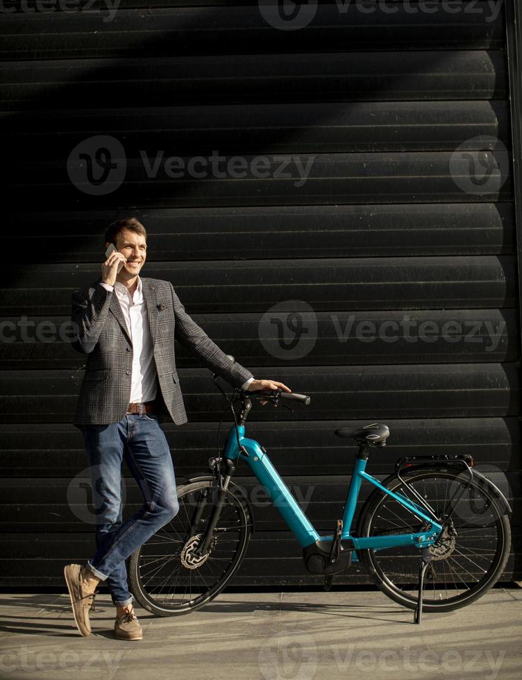 joven empresario de pie junto a la bicicleta eléctrica y usando un teléfono móvil foto