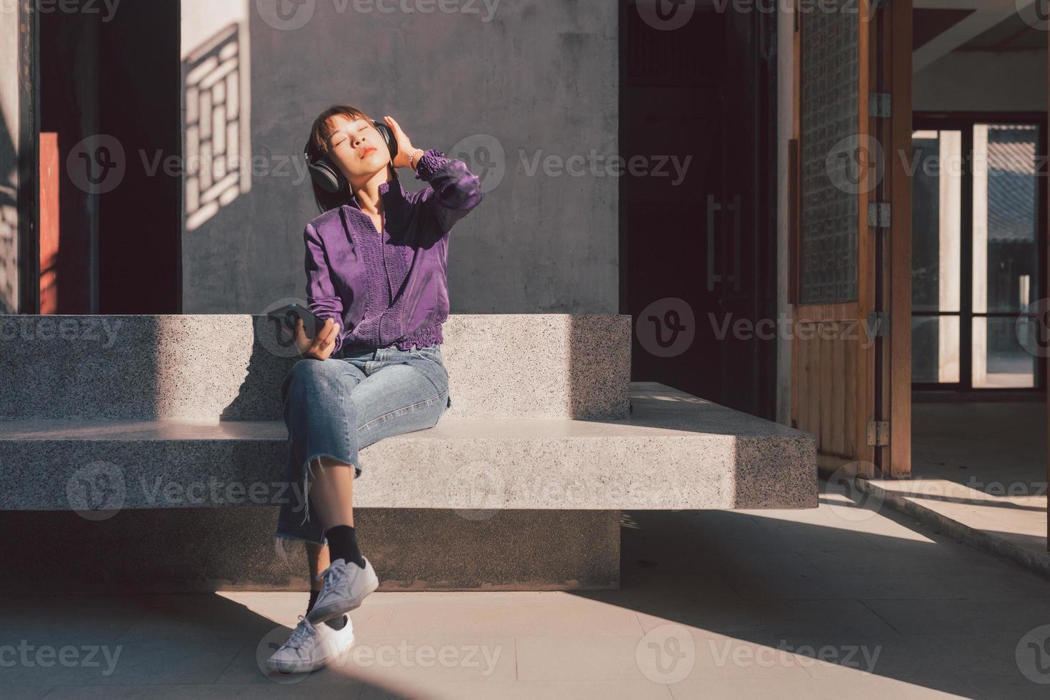 Feliz joven mujer asiática escuchando música con auriculares a través de un teléfono inteligente y divirtiéndose mientras está sentado al lado de la calle foto