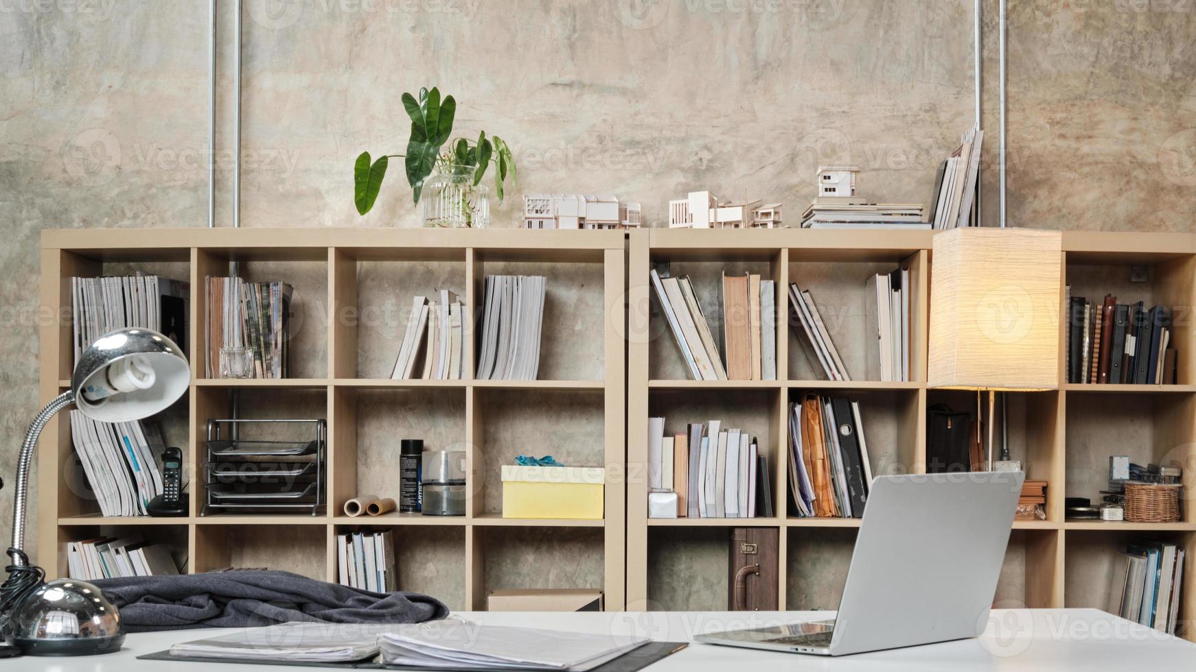 no hay personas en un lugar de trabajo informal, escritorio y silla de presidente en la oficina de arquitectura y diseño de interiores, modelos, libros, accesorios y equipos en la estantería decorados en estilo loft moderno. foto