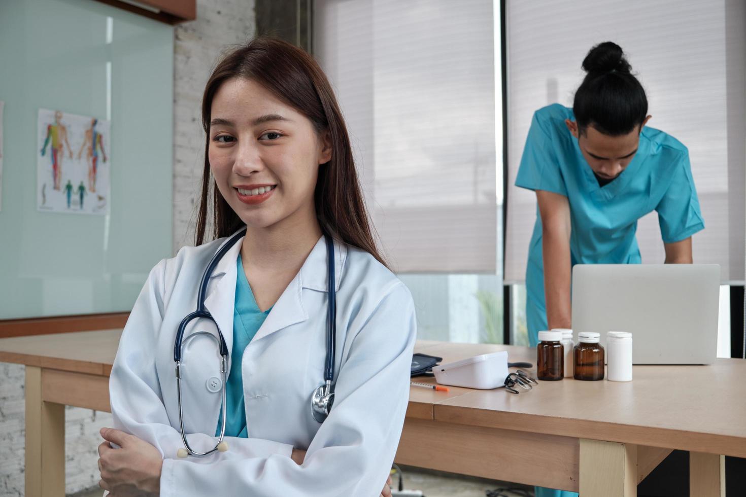 Retrato de hermosa doctora de origen étnico asiático en uniforme con estetoscopio. sonreír y mirar a la cámara en una clínica de hospital, pareja masculina trabajando detrás de ella, dos profesionales. foto