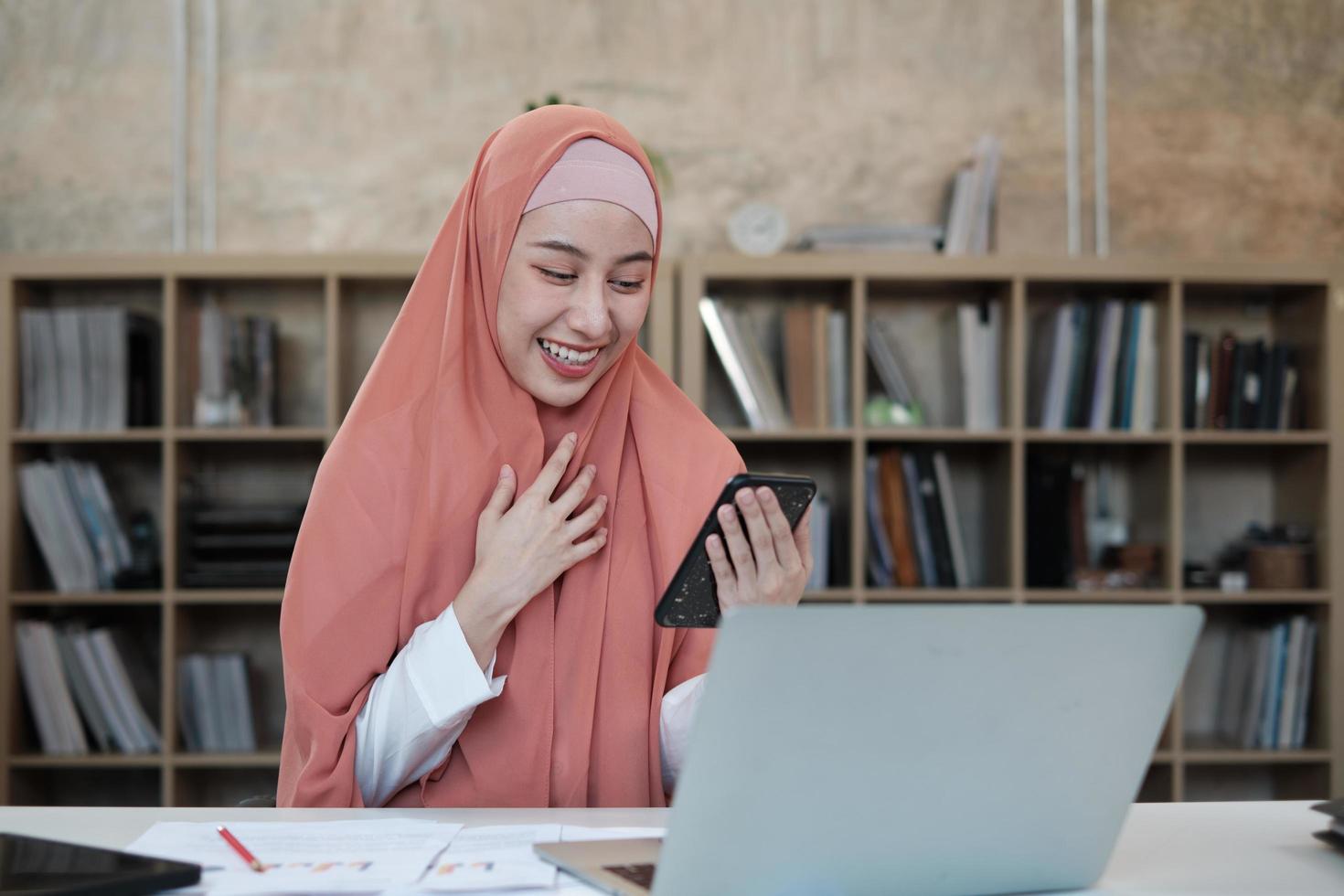 hermosa mujer de negocios que persona de sexo femenino islámico está hablando por la comunicación de su teléfono móvil y sonriendo feliz porque orden en línea de Internet en la computadora portátil, estilo de vida moderno, ocupación de comercio electrónico. foto