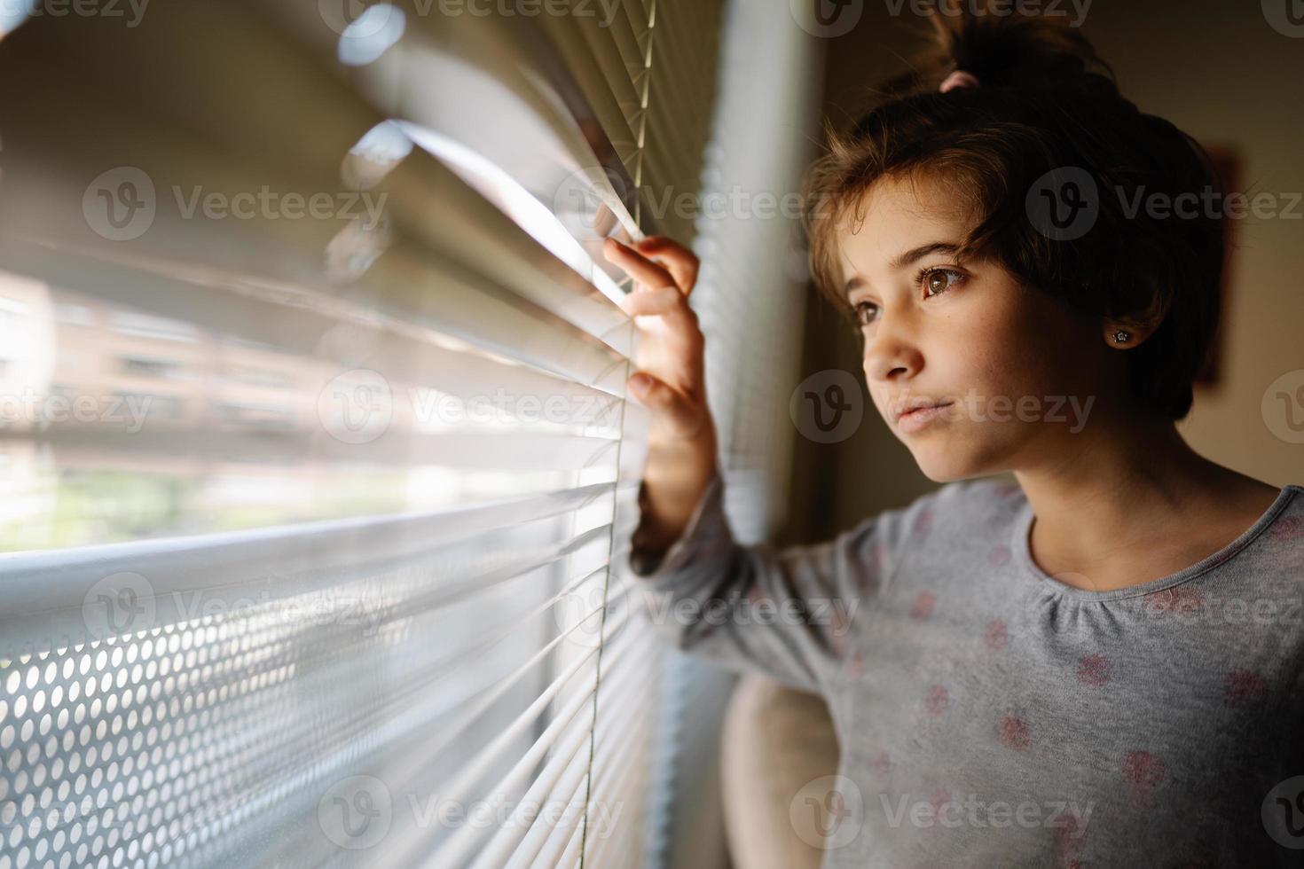 Niña de nueve años mirando por la ventana a través de las persianas. foto