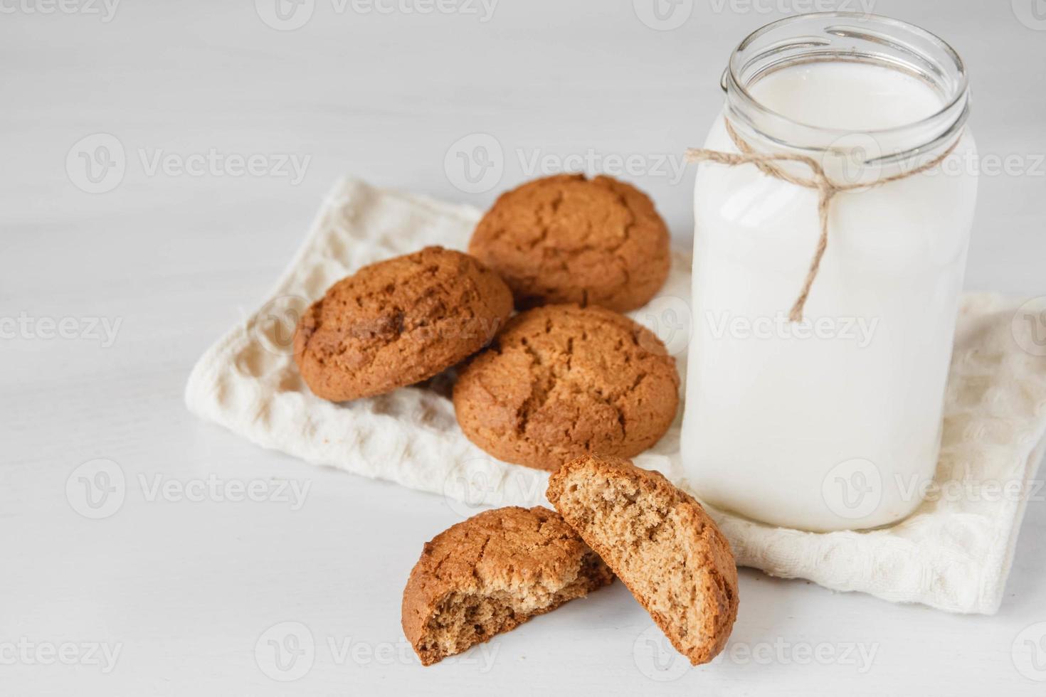 Milk in glass jar and oatmeal cookies near napkin on white table photo