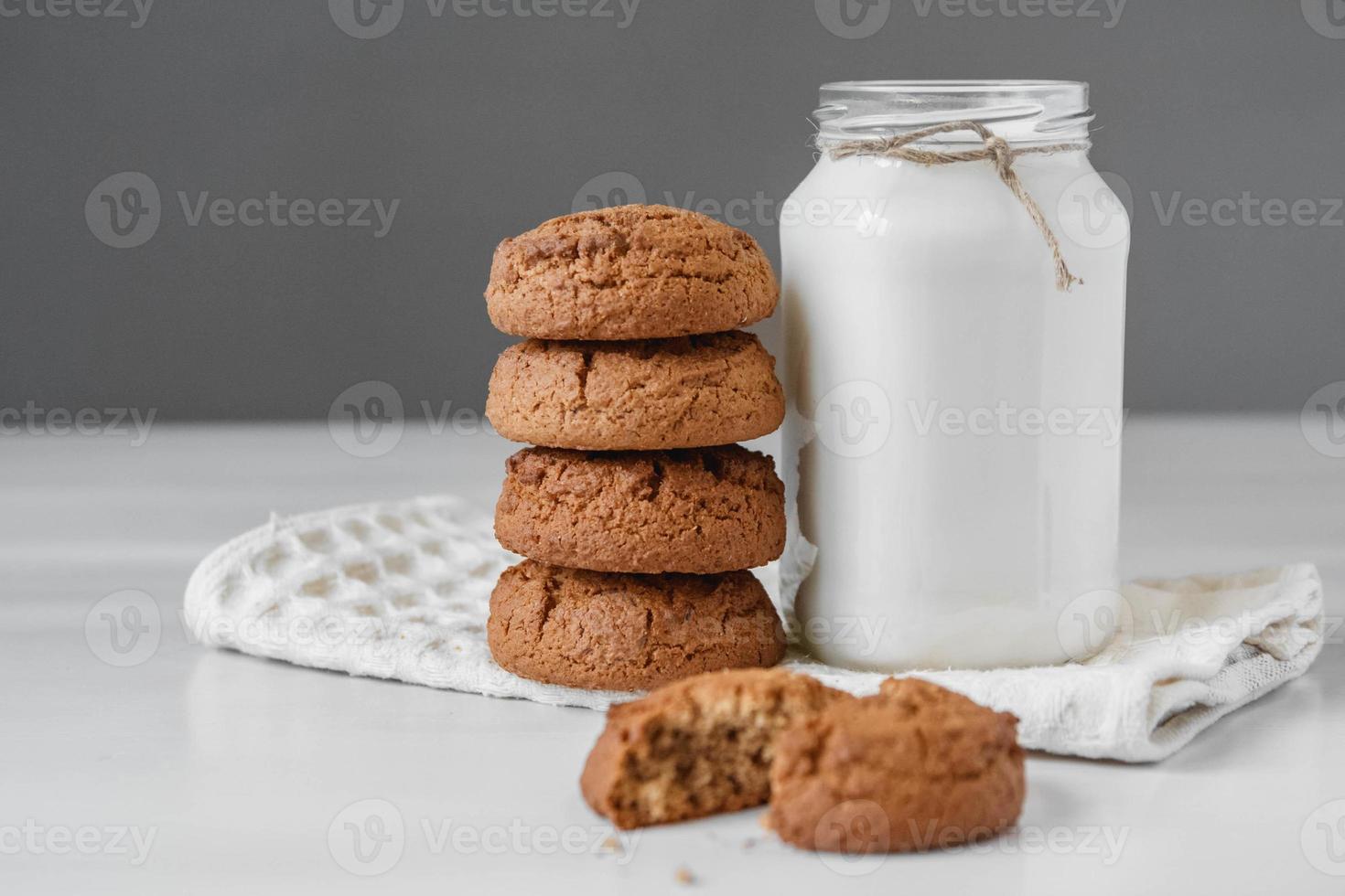 Milk in glass jar and oatmeal cookies near napkin on white table photo