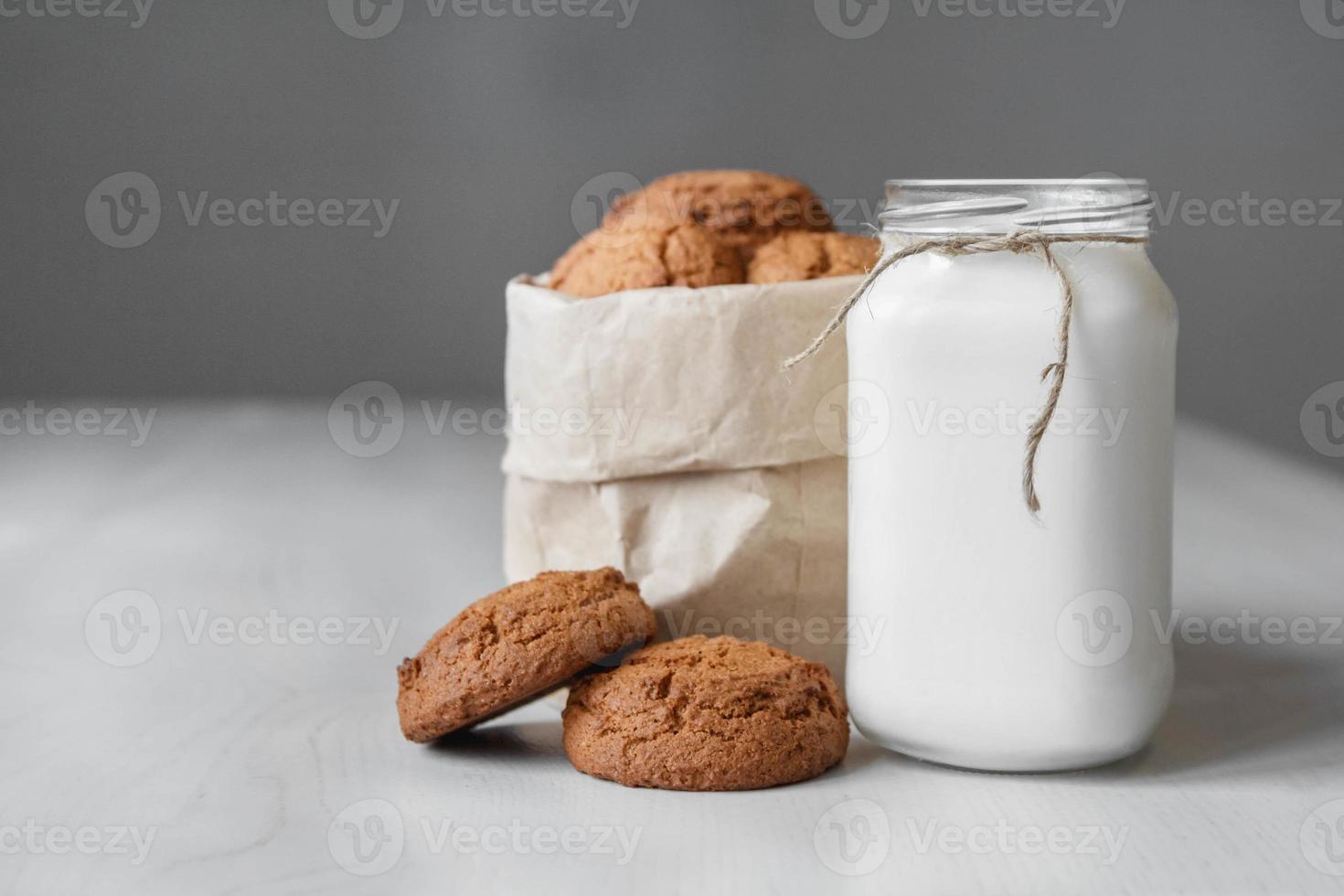 Leche en un frasco de vidrio y galletas de avena en una bolsa de papel sobre una mesa blanca foto