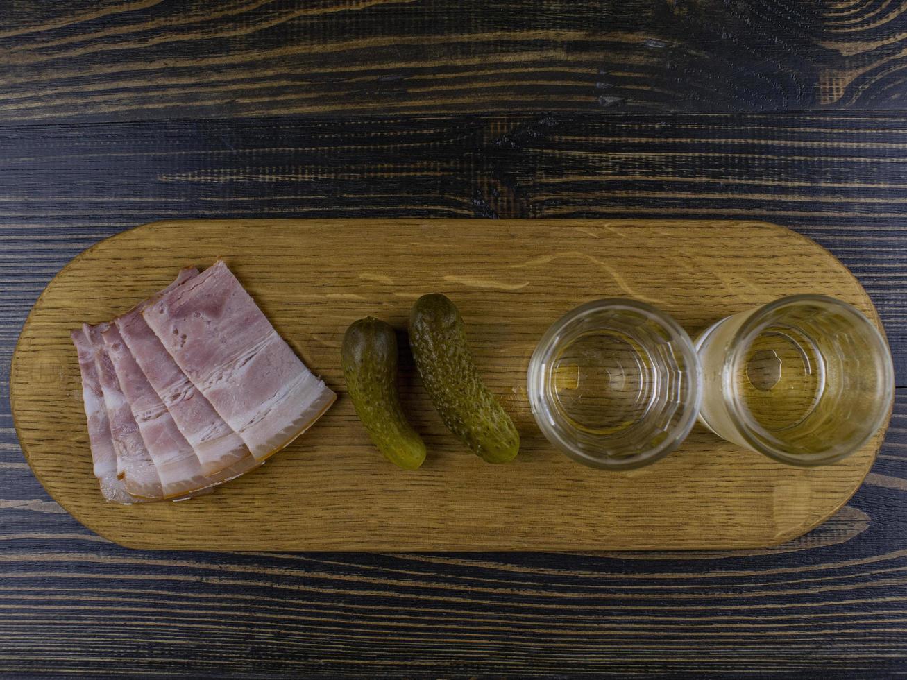 two misted glasses with cold vodka on a wooden board with bacon and pickled cucumbers. traditional russian snack photo