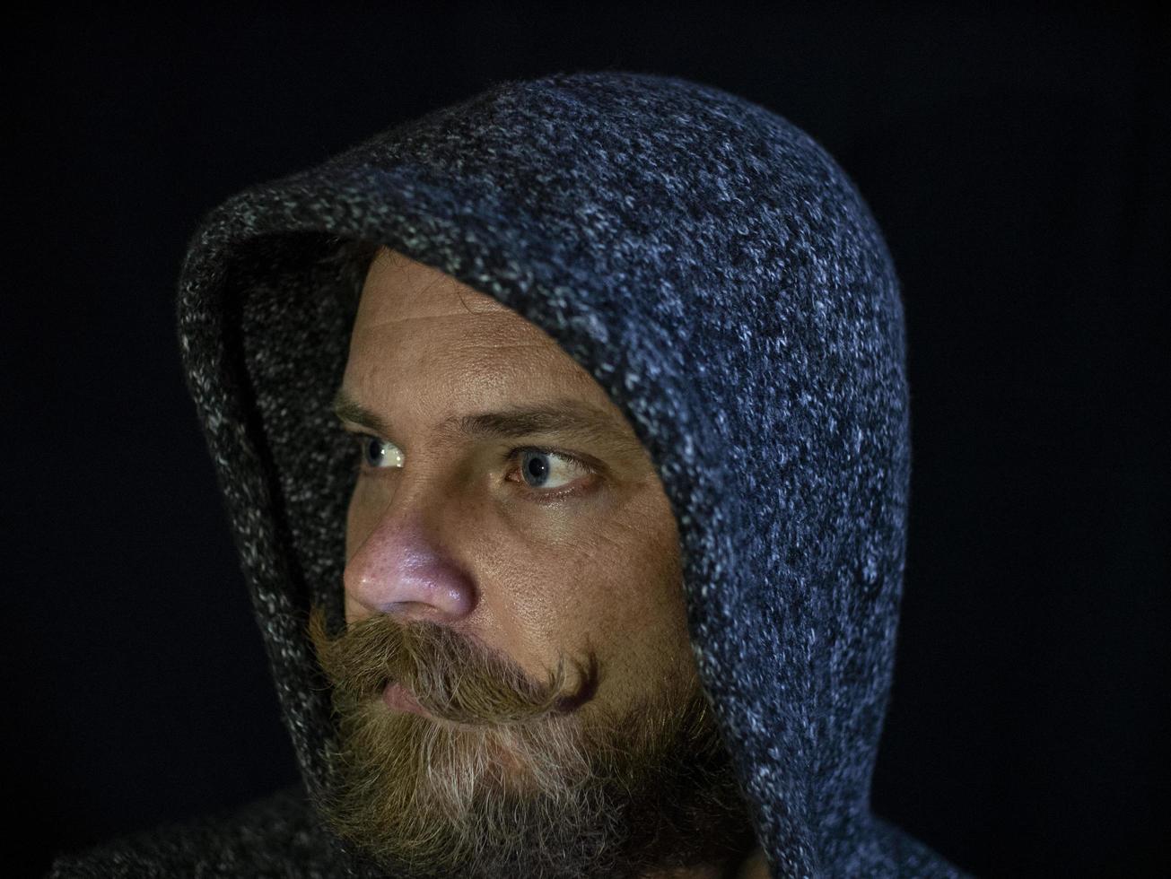 Retrato de un hombre con barba y bigote en el capó con un rostro serio sobre un fondo negro foto