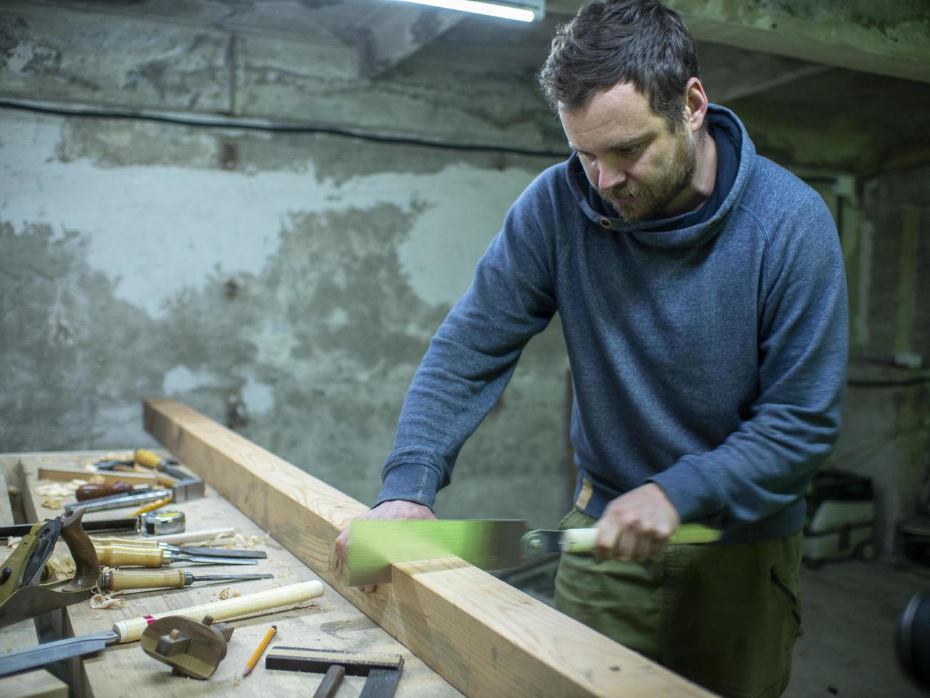 carpintero con barba aserrando una viga de madera con una sierra de mano. un carpintero aserrando un trozo de madera foto
