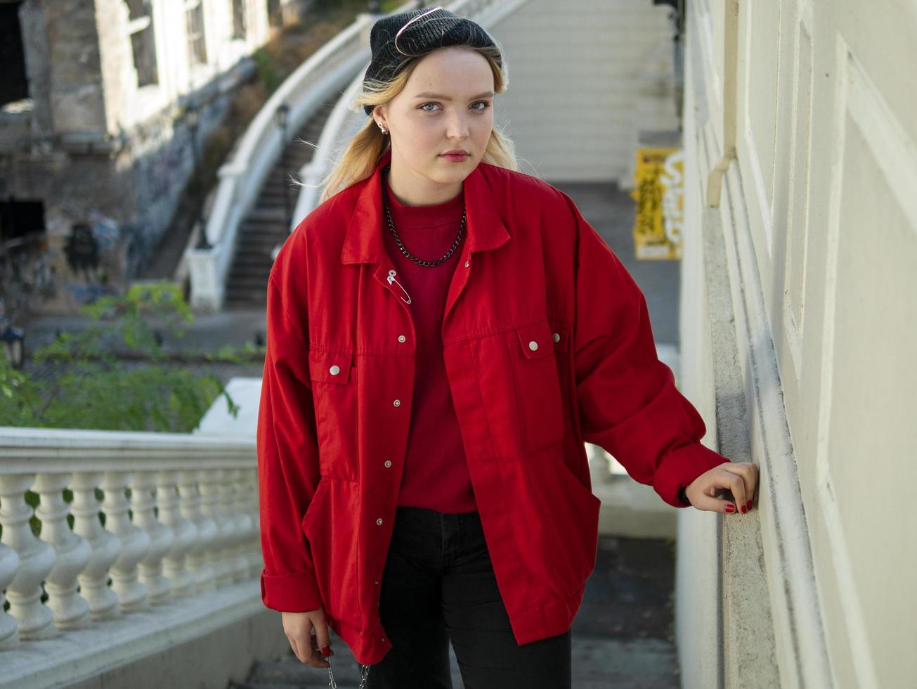Atractiva chica rubia hipster con un sombrero negro tejido y chaqueta roja está de pie en las escaleras debajo del puente foto