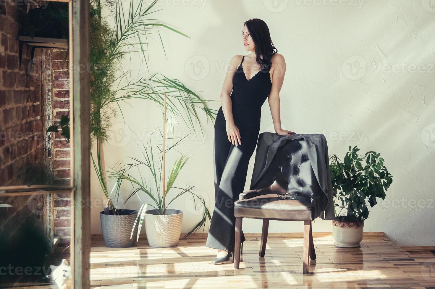 Woman dressed in black clothes standing near armchair in loft style interior photo