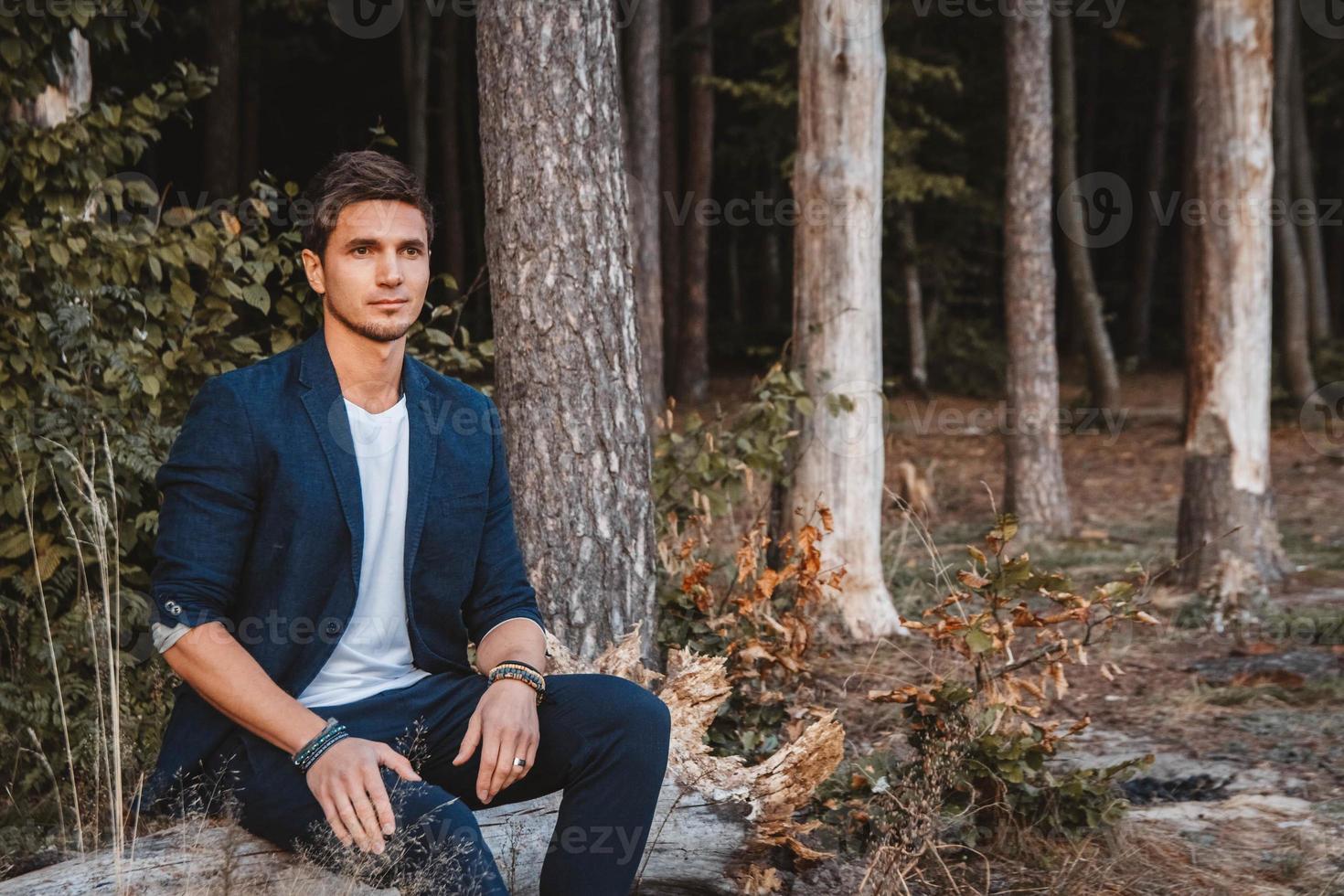 Man posing outdoors in forest sits on a log wearing checked jacket photo