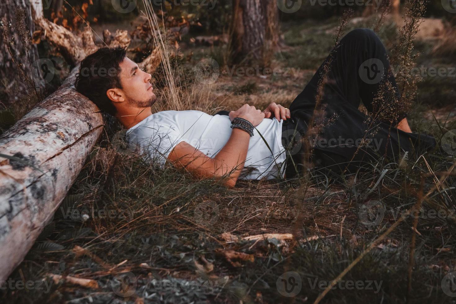Man lies on grass, resting his head on a log in middle of forest relaxing photo