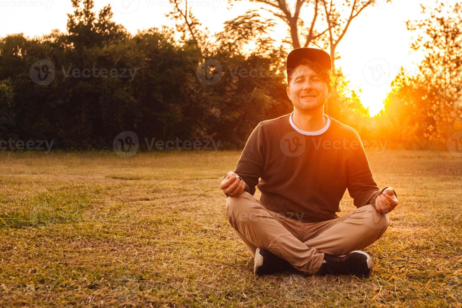 El hombre en una posición meditativa se sienta en un césped sobre un fondo de bosque y puesta de sol foto