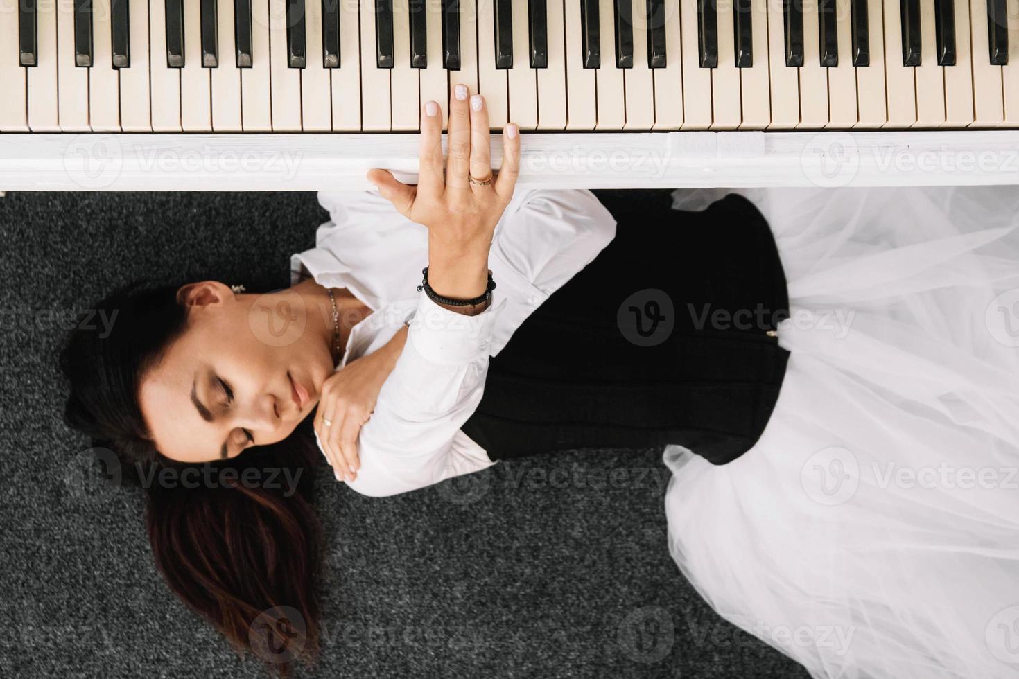 Woman dressed in a white dress with a black corset lies on the floor near white piano playing on the keys photo