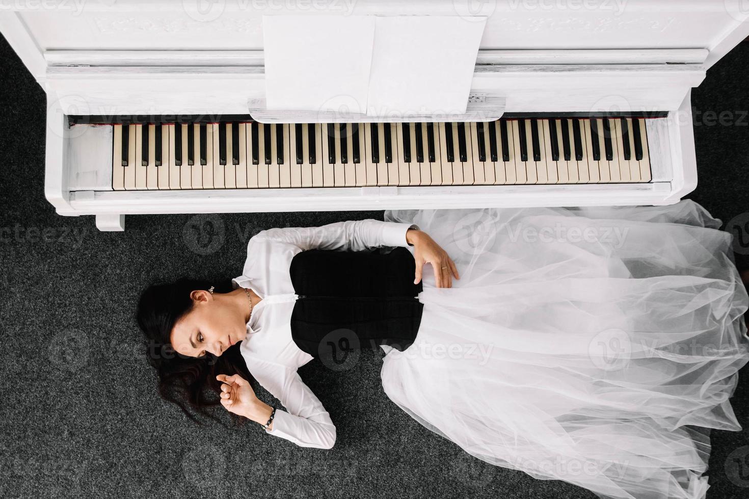 Beautiful woman dressed in a white dress with a black corset lies on the floor near white piano. Place for text or advertising. View from above photo