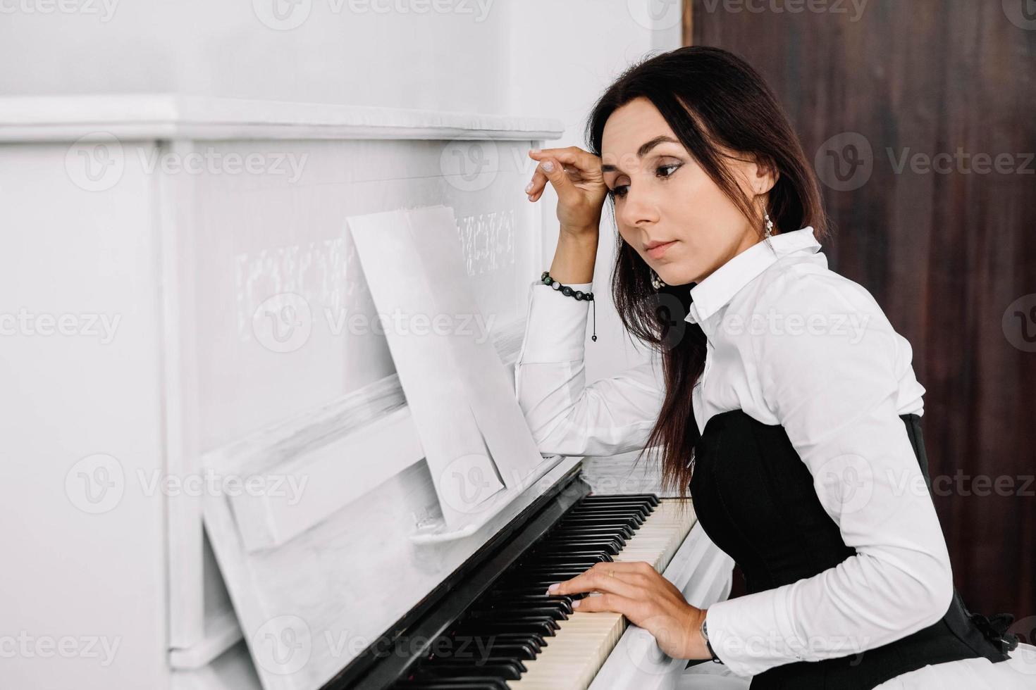 Beautiful woman dressed in white shirt looks into the musical notes and playing on white piano. Place for text or advertising photo