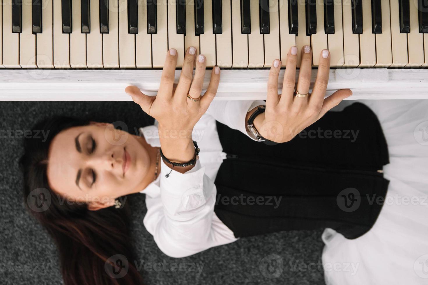 hermosa mujer vestida con un vestido blanco con un corsé negro yace en el suelo cerca de piano blanco tocando las teclas. lugar para texto o publicidad. vista desde arriba foto
