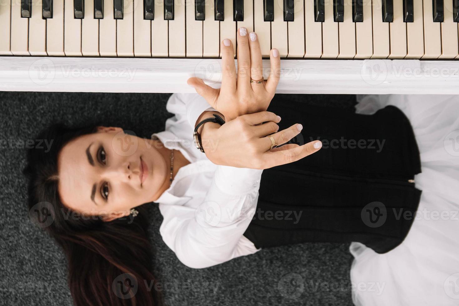 Beautiful woman dressed in a white dress with a black corset lies on the floor near white piano playing on the keys. Place for text or advertising. View from above photo