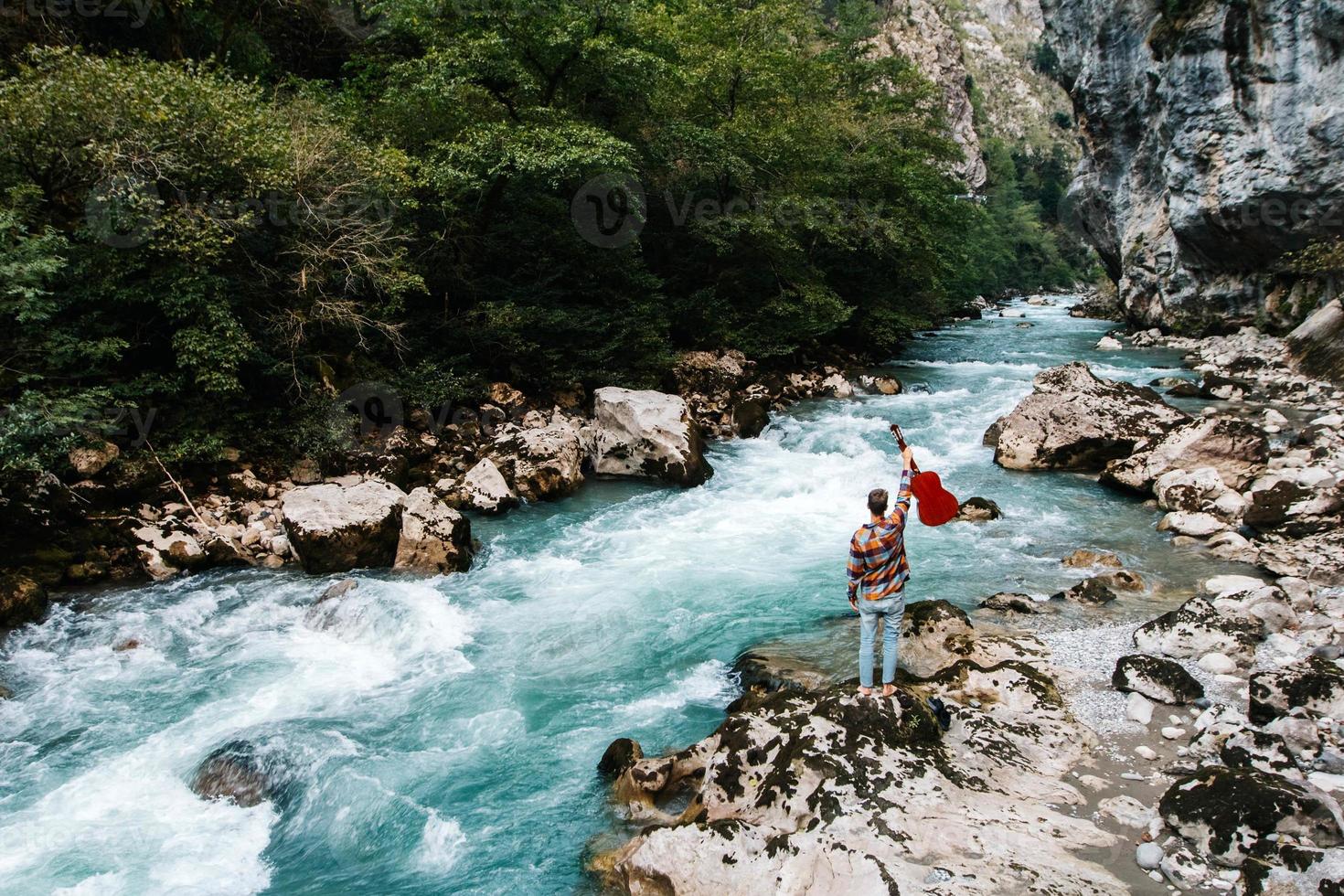 Man holding guitar standing on the bank of a mountain river on a background of rocks and forest. Handsome hippie style guitarist engrossed on music outdoors photo