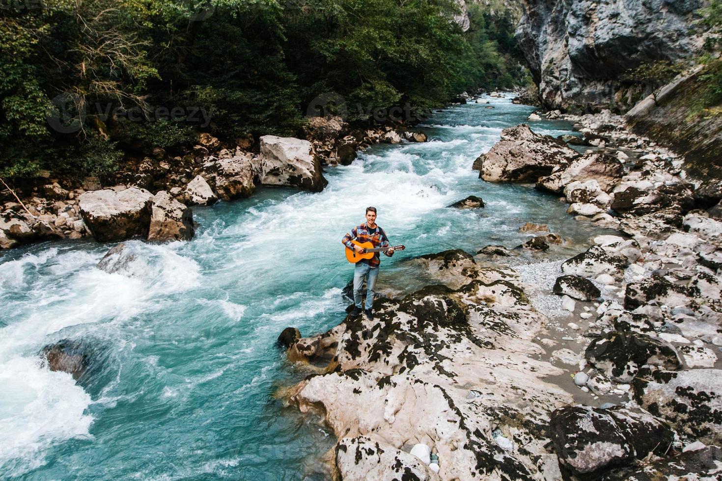 Man playing guitar standing on the bank of a mountain river on a background of rocks photo