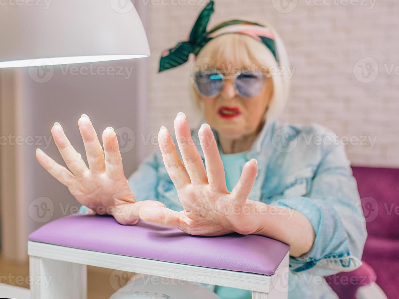 elderly stylish woman in blue sunglasses and jacket sitting at manicure salon unhappy with her nails. Treatment, care, anti age, manicure concept photo