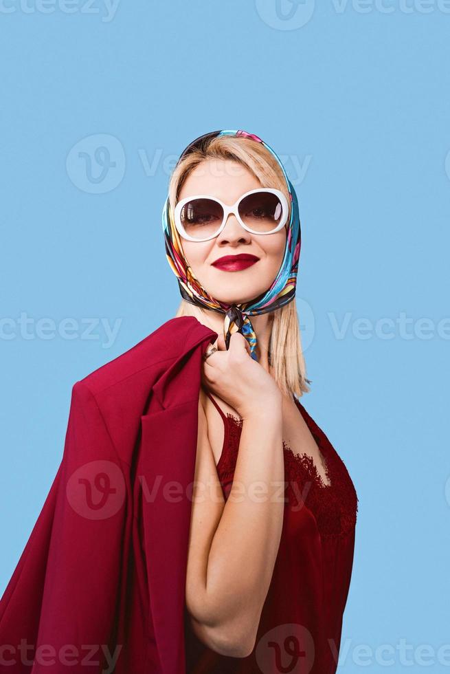 Elegante mujer rubia de fantasía alegre sonriente con maquillaje en pañuelo y gafas de sol sobre fondo azul. foto