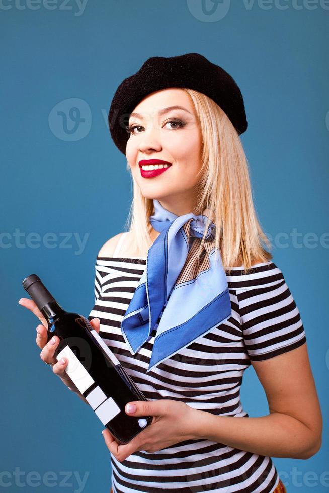 Retrato de una hermosa mujer francesa rubia en boina, bufanda, espalda y camisa blanca, con una botella de vino y pan baguette en sus brazos con la bandera francesa en el fondo foto