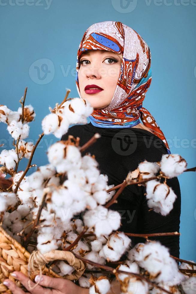 mujer oriental en colorido pañuelo con canasta con ramas de algodón sobre fondo azul. recolector de algodón foto