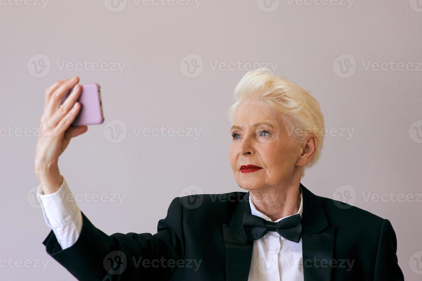 stylish mature senior woman in tuxedo with cellphone video calling or making selfie. Fun, party, style, lifestyle, business, technology, celebration concept photo