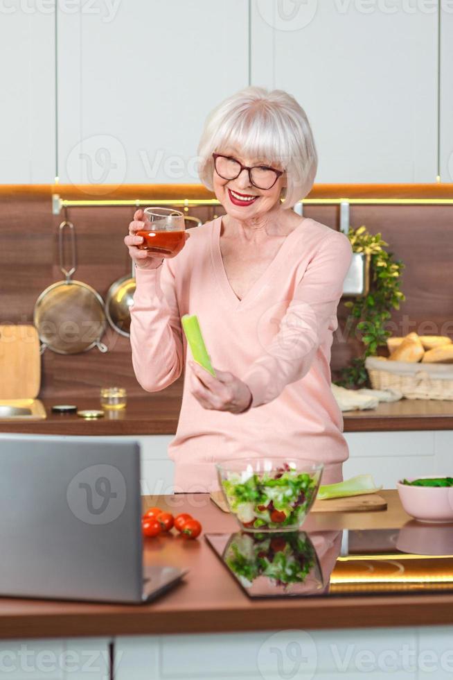 senior cheerful woman video blogger showing celery and tomato juice at her kitchen. Raw, vegetarian, diet, blogger, nutritionist kitchen concept photo